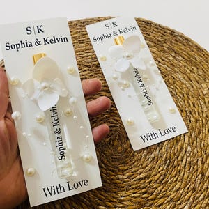 May include: Two white cards with perfume bottles, flower, and pearl embellishments. The cards are inscribed with "Sophia & Kelvin" and "With Love". The perfume bottles have gold caps and are decorated with white flowers and pearls.