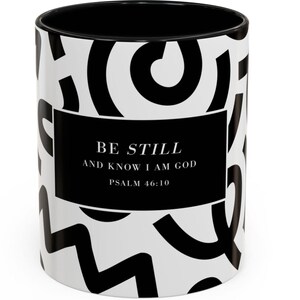 Könnte beinhalten: Weißer Keramikbecher mit schwarzem Innenraum und sich wiederholendem schwarzem, abstraktem Design. Ein schwarzes Rechteck zeigt den Text "BE STILL AND KNOW I AM GOD" und "PSALM 46:10".