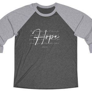 May include: A raglan baseball shirt with a gray body and gray sleeves. The shirt features the word "Hope" in a cursive font, along with a quote from Jeremiah 29:11. The text is in white.