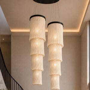 May include: Two tiered pendant lights featuring off-white fringe layers suspended from black circular ceiling mounts. Each fixture consists of four cascading cylindrical tiers of cream-colored corded fringe, illuminated from within by warm light.