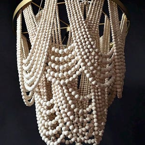 May include: A decorative chandelier featuring strands of off-white wooden beads cascading from a gold-colored metal frame. The beads are arranged in a layered, flowing pattern, creating a unique lighting fixture.