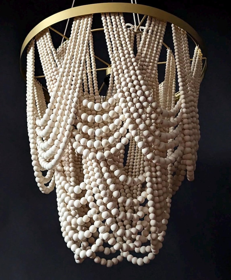 May include: A decorative chandelier featuring strands of off-white wooden beads cascading from a gold-colored metal frame. The beads are arranged in a layered, flowing pattern, creating a unique lighting fixture.