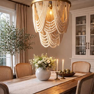 May include: A dining room featuring a wooden table with a light-colored runner, surrounded by woven-back chairs. A large, beaded chandelier hangs above the table. A vase of flowers and lit candles add to the ambiance. A cabinet is visible in the background.