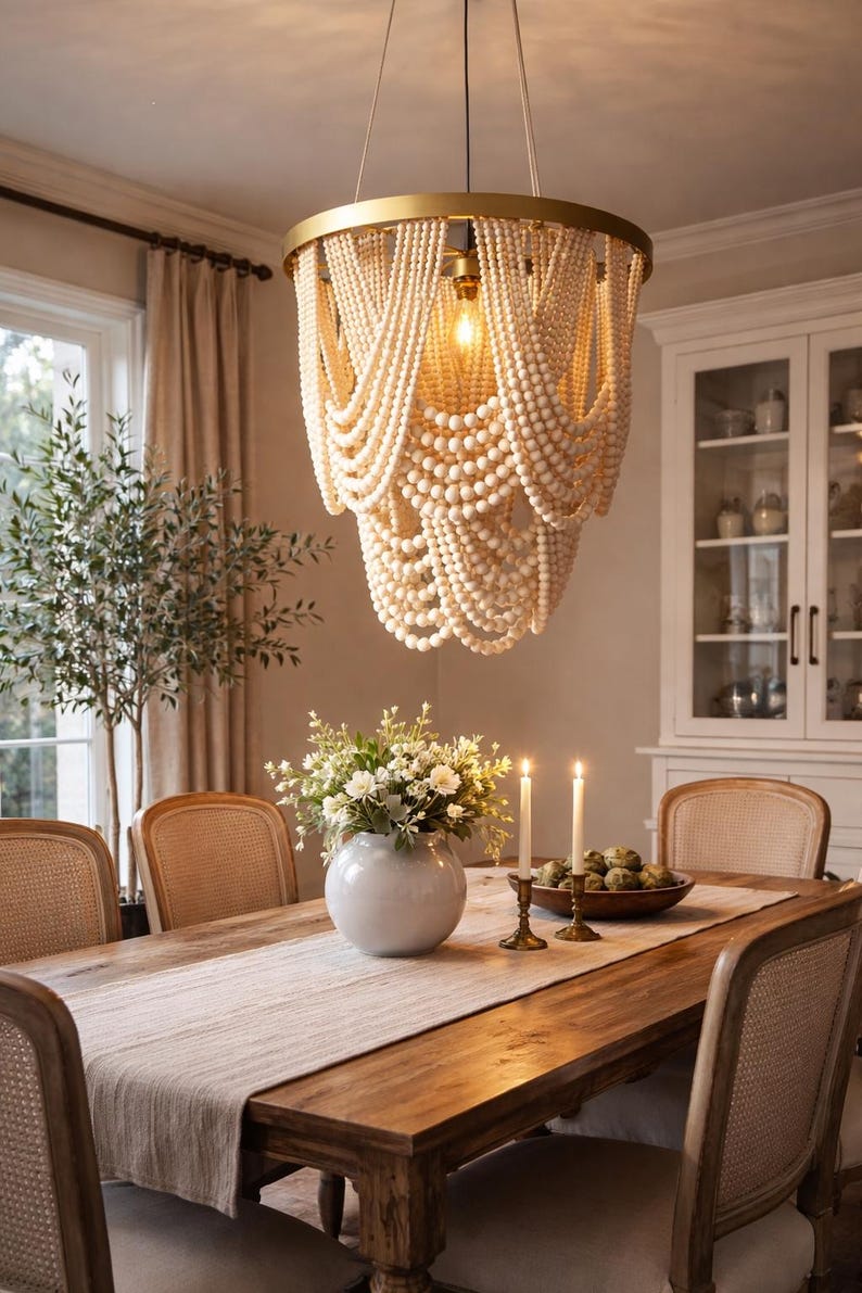 May include: A dining room featuring a wooden table with a light-colored runner, surrounded by woven-back chairs. A large, beaded chandelier hangs above the table. A vase of flowers and lit candles add to the ambiance. A cabinet is visible in the background.