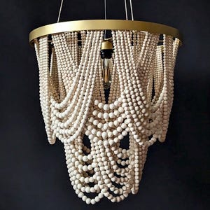 Puede incluir: Lámpara colgante decorativa con un anillo metálico dorado y filas de cuentas de madera blanquecinas. Las cuentas están dispuestas en hebras fluidas. Una única bombilla cuelga del centro. Diseño elegante y decorativo.