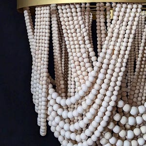 May include: A decorative chandelier featuring strands of light-colored wooden beads cascading from a gold-colored metal ring. The beads are arranged in a flowing, layered pattern, creating a textured and elegant design. The chandelier hangs against a dark background.
