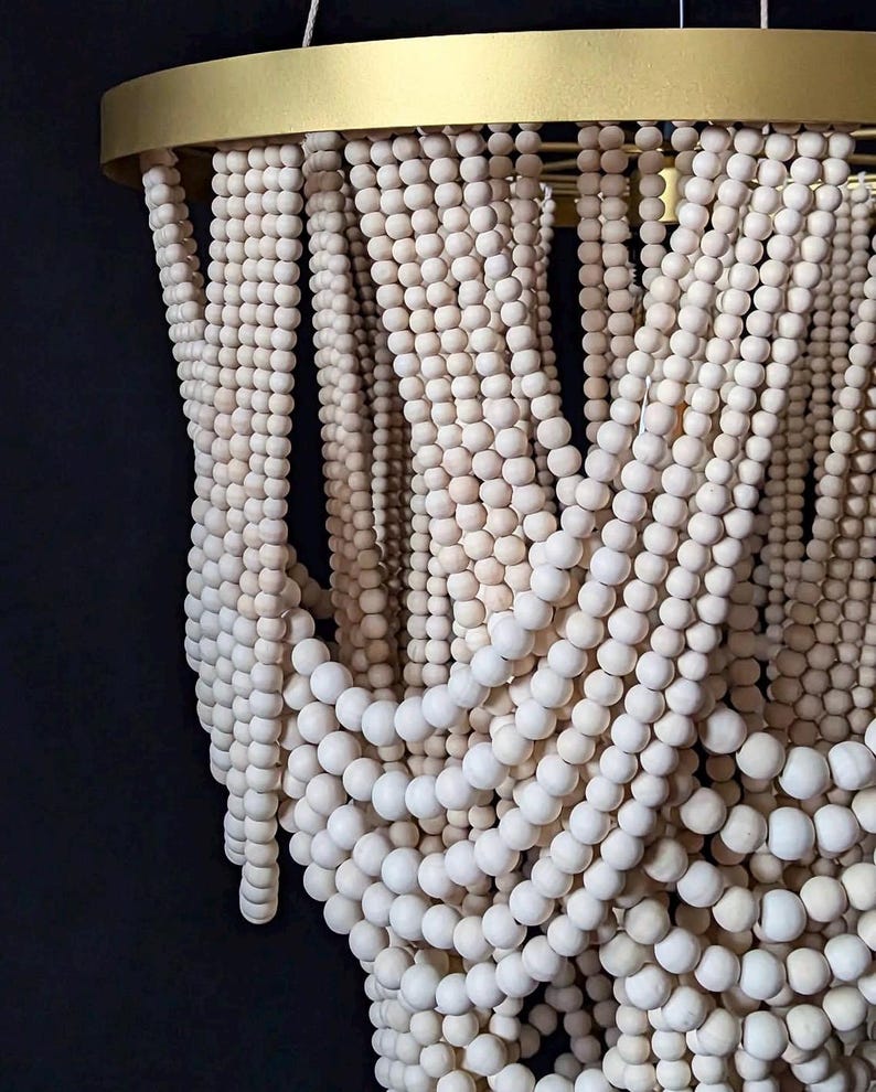 May include: A decorative chandelier featuring strands of light-colored wooden beads cascading from a gold-colored metal ring. The beads are arranged in a flowing, layered pattern, creating a textured and elegant design. The chandelier hangs against a dark background.