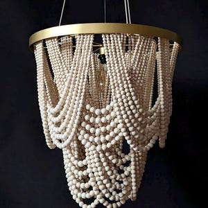 May include: A decorative chandelier with a gold-colored metal frame and cascading strands of white wooden beads. The light fixture hangs from thin wires and features a single exposed lightbulb. The design is elegant and suitable for interior decor.
