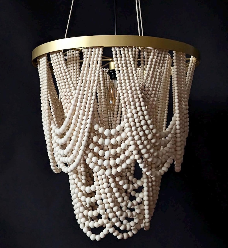 May include: A decorative chandelier with a gold-colored metal frame and cascading strands of white wooden beads. The light fixture hangs from thin wires and features a single exposed lightbulb. The design is elegant and suitable for interior decor.