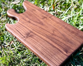 Puede incluir: Una tabla de cortar rectangular de madera con asa. La tabla es de un color marrón oscuro intenso y tiene un acabado liso y pulido. El asa es curva y tiene un diseño recortado. La tabla está sobre hierba verde.