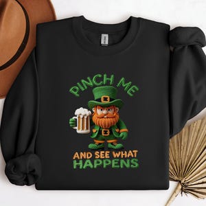 Op de afbeelding: Zwart sweatshirt met een groene en oranje cartoon kabouter die een bierpul vasthoudt. De tekst "PINCH ME AND SEE WHAT HAPPENS" is geborduurd in groen en oranje.