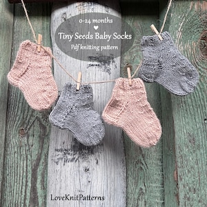 May include: Four knitted baby socks in shades of pink and grey are hung on a washing line with wooden pegs. The socks are labelled "Tiny Seeds Baby Socks" and are designed for babies aged 0-24 months. The background is a weathered wooden surface.
