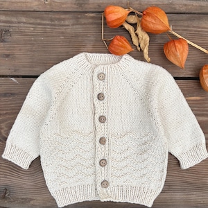 May include: Cream-colored knitted baby cardigan with long sleeves and a round neckline. The sweater features a chevron pattern on the lower front and is fastened with five wooden buttons. The background includes dried orange flowers and a wooden surface.