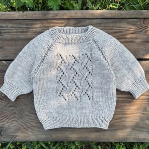 May include: A light gray knitted sweater with a diamond pattern on the front. The sweater has long sleeves and a ribbed hem and cuffs. The neckline is also ribbed. The sweater is made of a soft, textured yarn.