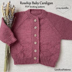 May include: A dusty rose knitted baby cardigan with long sleeves, a round neckline, and six light-colored wooden buttons down the front. The text Rosehip Baby Cardigan PDF knitting pattern and 0-24 months is visible above the garment.