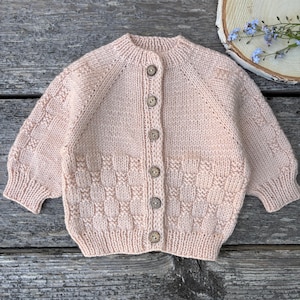 May include: A pale pink long-sleeve knit cardigan featuring a round neckline, ribbed cuffs, and a vertical row of six brown wooden buttons down the centre. The lower portion of the garment and the sleeves display a textured basketweave pattern.