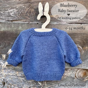 May include: A blue knitted baby sweater, displayed on a wooden hanger shaped like bunny ears. The sweater has long sleeves and a ribbed hem and cuffs. Text on the image reads "Blueberry Baby Sweater" and "0-24 months".