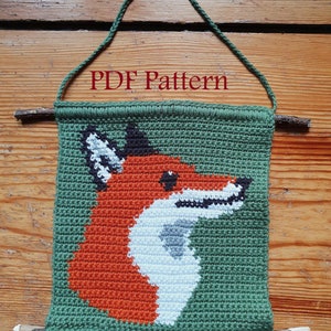 Könnte beinhalten: Ein gehäkelter Wandbehang mit einem Fuchs in orange, weiß und schwarzen Details. Der Fuchs befindet sich auf einem grünen Hintergrund. Der Behang ist an zwei Holzstäben mit einer Schlaufe aus Seil oben befestigt. Der Text "PDF Pattern" ist oben im Bild sichtbar.