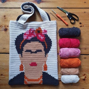 Könnte beinhalten: Eine gehäkelte Einkaufstasche mit einem Porträt von Frida Kahlo. Die Tasche ist aus weißem, schwarzem, orangefarbenem, rosa und rotem Garn gefertigt. Die Tasche wird von Garnknäueln in verschiedenen Farben begleitet.