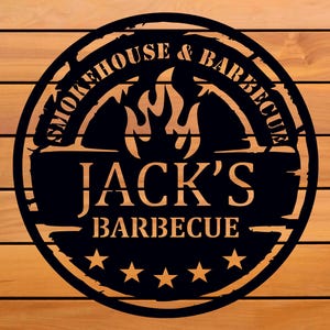 Op de afbeelding: Zwart metalen bord met de tekst "Smokehouse & Barbecue" en "Jack's Barbecue" in een cirkelvormig ontwerp. Het bord heeft een vlammenafbeelding en vijf sterren, tegen een houten achtergrond.
