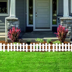 Puede incluir: Una valla blanca bordea un césped verde vibrante frente a una casa. La casa tiene un exterior gris, una puerta blanca y detalles de piedra. Arbustos de color púrpura rojizo y otra vegetación añaden color.