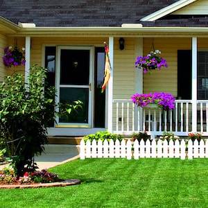 May include: Exterior view of a house with a yellow facade, white trim, and a dark roof. The porch has hanging baskets of purple flowers and a white picket fence borders the green lawn.
