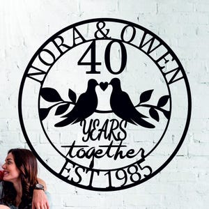 May include: Black metal wall art celebrating a 40th wedding anniversary. The design features the names "Nora & Owen", the number "40", two doves, and the words "Years together EST 1985" in a circular frame.