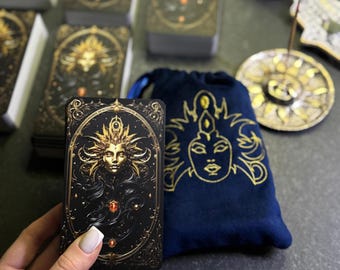 Puede incluir: Una carta de tarot dorada y negra con el rostro de una mujer rodeado por un halo dorado. La carta se sostiene en una mano y un bolso de terciopelo azul con bordados dorados está en el fondo.