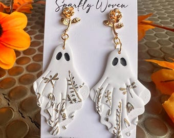May include: White ghost-shaped earrings with black eyes and gold floral detailing. The earrings are suspended from gold-coloured rose studs. The card behind the earrings reads "Sparkly Woven".
