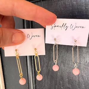 May include: Two pairs of earrings are displayed on pink cards. One pair has gold-colored rectangular frames with pink beads. The other pair has silver-colored rectangular frames with pink beads. The cards have the text "Sparkly Woven" and "-Woven".