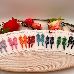 May include: A collection of colorful, handmade earrings displayed on white cards. The earrings are in various colors, including lavender, pink, orange, red, teal, navy, green, and black. Each pair features a unique design. The cards are arranged on a wooden tray with floral accents.