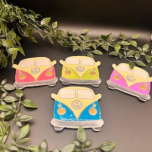 May include: A collection of five colorful, hand-drawn coasters shaped like vintage camper vans. Each coaster features a different color scheme, including red, green, pink, blue, and yellow, with a white outline and a VW logo.