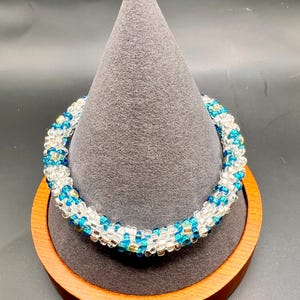 May include: A beaded bracelet with a spiral design in shades of blue, white, and gold. The bracelet is displayed on a wooden stand.
