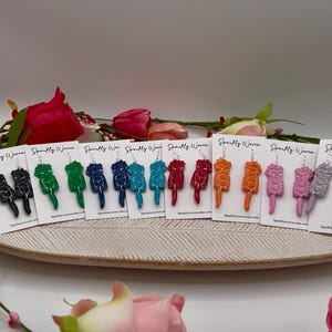 May include: A collection of colorful, handmade earrings displayed on a wooden tray. The earrings are in various colors, including black, green, blue, red, orange, pink, and gray. Each earring has a unique, abstract design and is attached to a small white card.