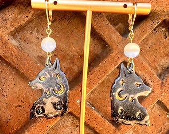 May include: A pair of gold dangle earrings featuring a black and gold wolf silhouette with moon and star details. Each earring has a white pearl bead.