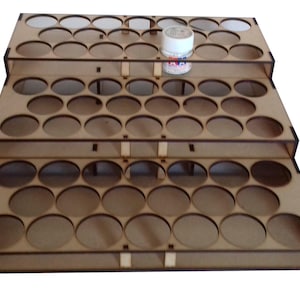 May include: A three-tiered wooden paint rack with 36 circular slots for holding paint bottles. The rack is made of light brown wood and has a white plastic bottle on the top tier.
