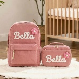 May include: A pink corduroy backpack and matching lunch bag set. The backpack and lunch bag have the name "Bella" embroidered in white script, with a pink flower accent. The set is designed for children.