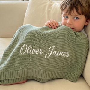 May include: A sage green knitted blanket with the embroidered name "Oliver James" in white script. The blanket is draped over a person sitting on a cream-coloured sofa. The blanket appears soft and cosy.