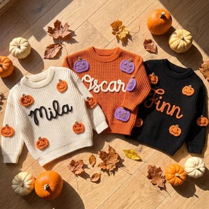 Peut inclure: Trois pulls en tricot à manches longues de couleur crème, orange et noire. Le pull crème porte le nom Mila, le pull orange porte le nom Oscar avec des appliqués de citrouilles violettes, et le pull noir porte le nom Finn.