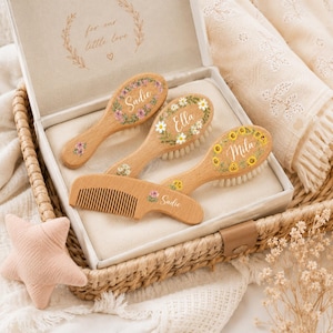 May include: A wooden baby hairbrush and comb set with floral designs and personalized names. The set includes three brushes and a comb, each with a different floral pattern and name. The box reads "for our little love".