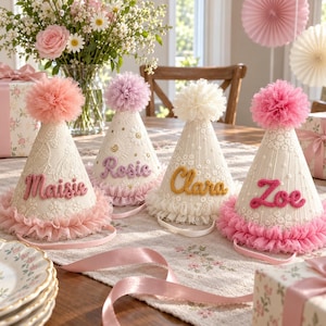 May include: Four cone-shaped party hats featuring cream-colored fabric with floral lace patterns, ruffled tulle trim at the base, and a large tulle pom-pom on top. Each hat displays a name in cursive yarn lettering: Maisie, Rosie, Clara, and Zoe.