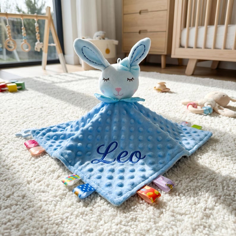 Puede incluir: Una manta de seguridad azul para beb&eacute; con una cabeza de conejo de peluche. La manta tiene una superficie texturizada y el nombre "Leo" bordado. Las etiquetas coloridas est&aacute;n cosidas alrededor del borde. Un art&iacute;culo reconfortante para beb&eacute;s.