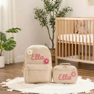 以下が含まれることがあります： ベージュのコーデュロイバックパックと、お揃いのランチバッグ。どちらのアイテムにも、ピンクのスクリプトで「Ella」という名前が書かれており、花のアップリケが飾られています。バックパックにはフロントポケットと上部のハンドルが付いています。ランチバッグには上部のハンドルとジッパー開閉部が付いています。
