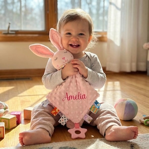 Puede incluir: Una manta de seguridad rosa para beb&eacute; con una cabeza de conejito y el nombre "Amelia" bordado. La manta tiene un mordedor en forma de estrella y etiquetas coloridas. El conejito tiene orejas rosas y una cara sonriente.