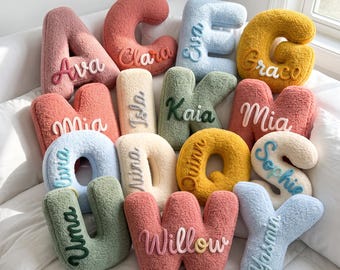 Peut inclure: Une collection de coussins en forme de lettres, texturés et moelleux, dans diverses couleurs pastel et douces. Chaque coussin présente un prénom brodé en écriture cursive sur le devant. Les lettres incluent A, C, E, G, M, I, K, M, U, O, Q, S, U, W et Y, avec des prénoms comme Ava, Clara, Eva, Grace, Mia, Isla, Kaia, Mia, Liva, Nina, Quinn, Sophie, Uma, Willow et Yasmin.