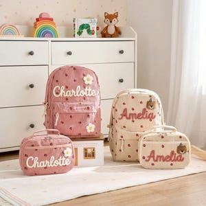 Könnte beinhalten: Rosa und beige Cordrucksäcke und Lunchpakete mit Erdbeer- und Blumendetails. Die Rucksäcke tragen die Namen "Charlotte" und "Amelia" aufgestickt. Eine passende Lunchtasche ist ebenfalls abgebildet.