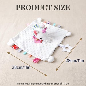 Puede incluir: Una manta de seguridad para beb&eacute; blanca con una cabeza de unicornio de peluche y cintas de colores. La manta mide 28 cm por 28 cm y tiene una superficie texturizada. Un mordedor en forma de estrella est&aacute; adjunto.
