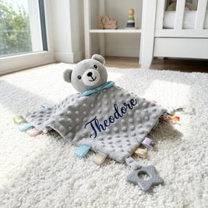 Puede incluir: Una manta de seguridad gris para beb&eacute; con una cabeza de oso de peluche y el nombre "Theodore" bordado en azul. La manta tiene una superficie texturizada, cintas de colores y un mordedor en forma de estrella.