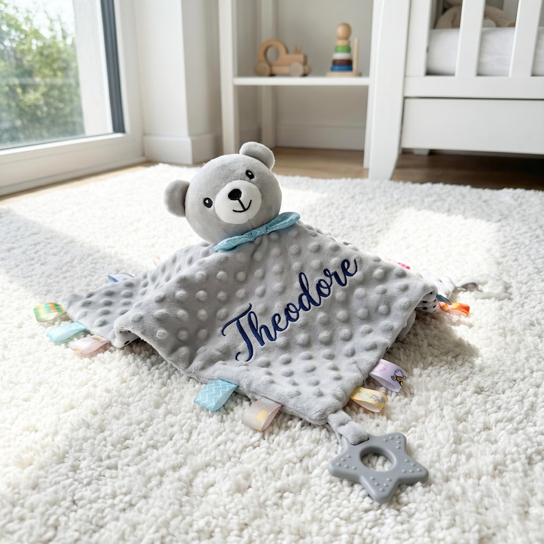 Puede incluir: Una manta de seguridad gris para beb&eacute; con una cabeza de oso de peluche y el nombre "Theodore" bordado en azul. La manta tiene una superficie texturizada, cintas de colores y un mordedor en forma de estrella.