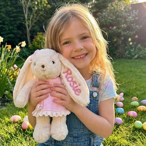 May include: A plush bunny rabbit toy in a pink dress with the name "Rosie" embroidered on one ear. The bunny has long, floppy ears and is being held by a smiling child. Easter eggs are scattered on the grass in the background.